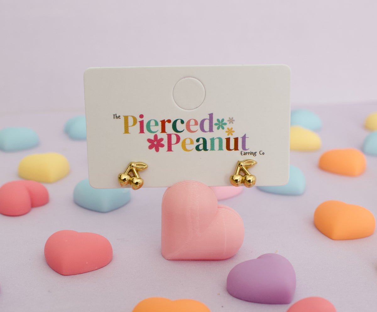 Hypoallergenic Gold cherry stud screwback earrings for kids on a white display card from 'The Pierced Peanut Earring Co', set against a pastel pink background decorated with colorful heart-shaped candies.