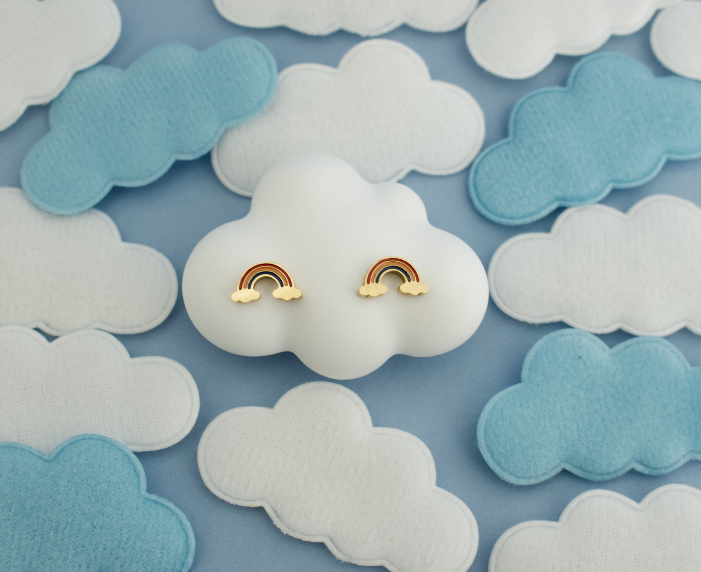 A pair of small kids hypoallergenic rainbow stud earrings with gold-tone metal and pastel rainbow enamel, displayed on a white cloud-shaped stand against a blue background with white and light blue felt clouds.