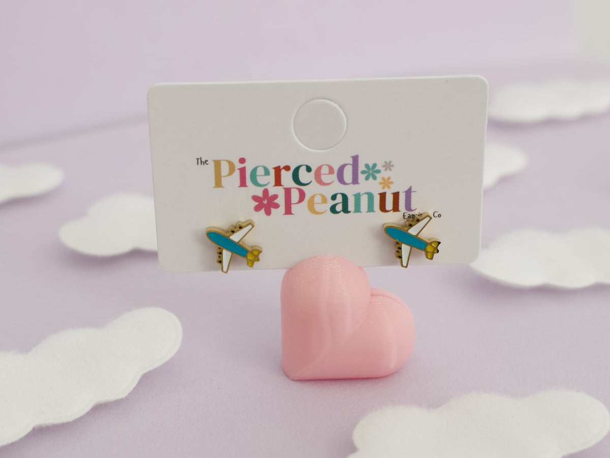 A pair of small hypoallergenic kids airplane screwback stud earrings displayed on a white backing card that reads 'The Pierced Peanut.' The earrings have turquoise-blue and gold wings with small pink details.