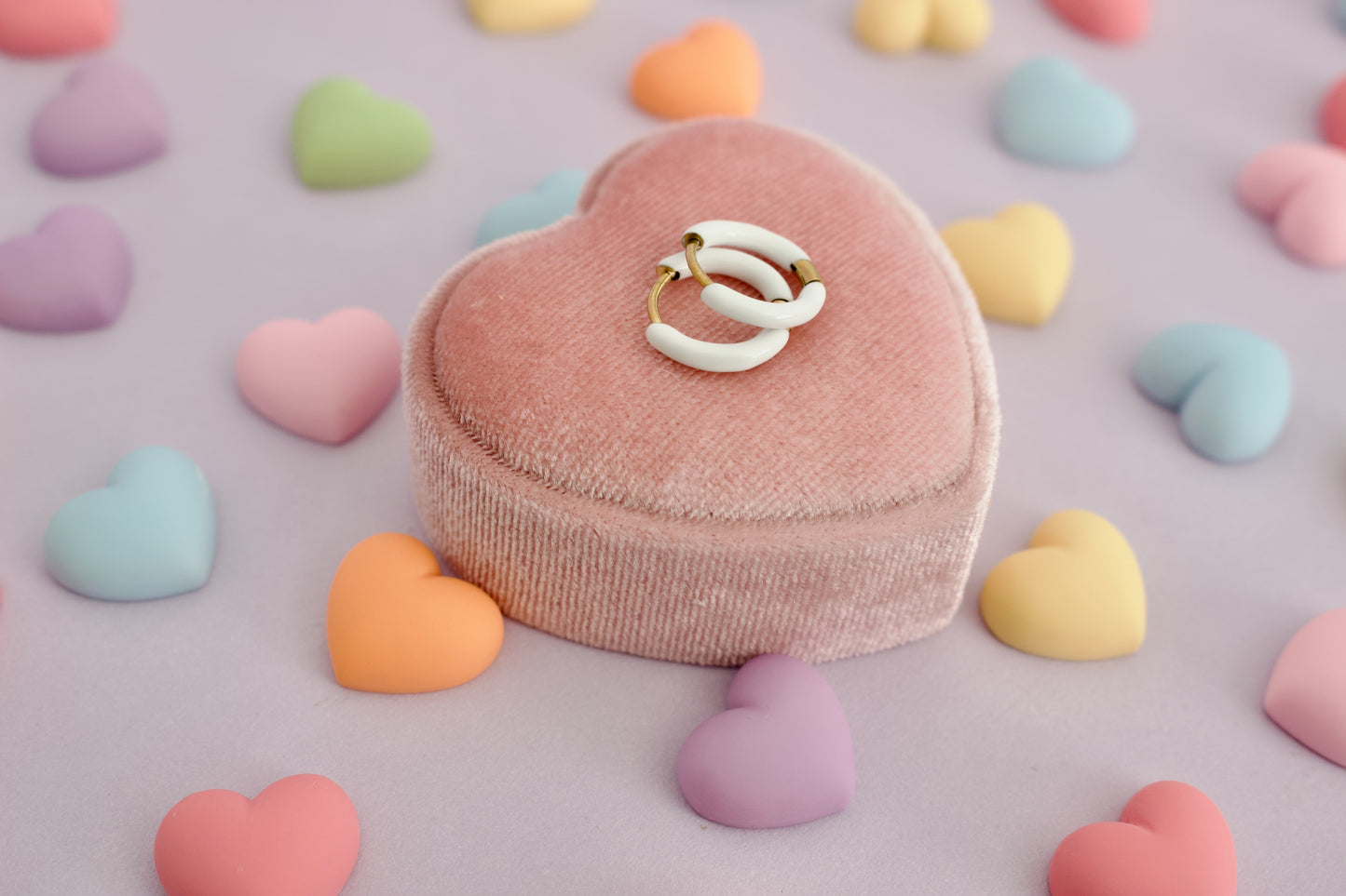 A Pair of kids hypoallergenic gold and white enamel huggie hoop earrings sitting on a pink heart-shaped velvet ring box, surrounded by pastel candy hearts on a lavender background. Earrings are from The Pierced Peanut Earring Co.