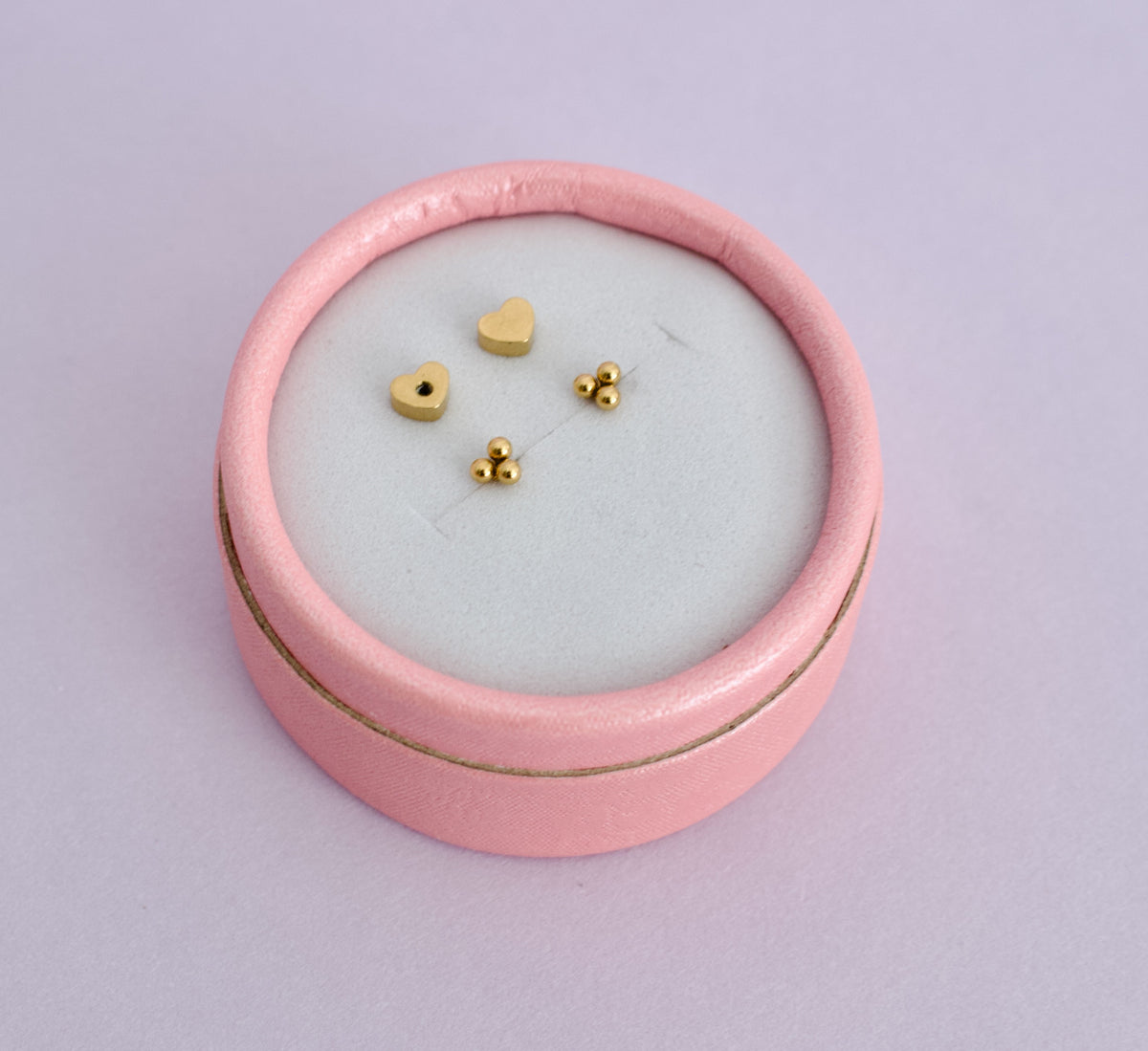 A pair of kids hypoallergenic gold trinity ball screwback stud earrings with heart shaped backs. Displayed inside a pale pink ring box from the brand The Pierced Peanut Earring Co based in New Zealand.