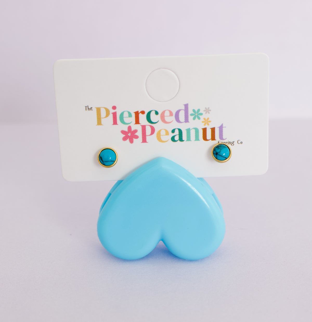 A pair of gold-framed stud earrings with round turquoise blue cabochon stones, displayed on a white backing card that reads 'The Pierced Peanut Earring Co.' with a light blue heart-shaped prop in front.