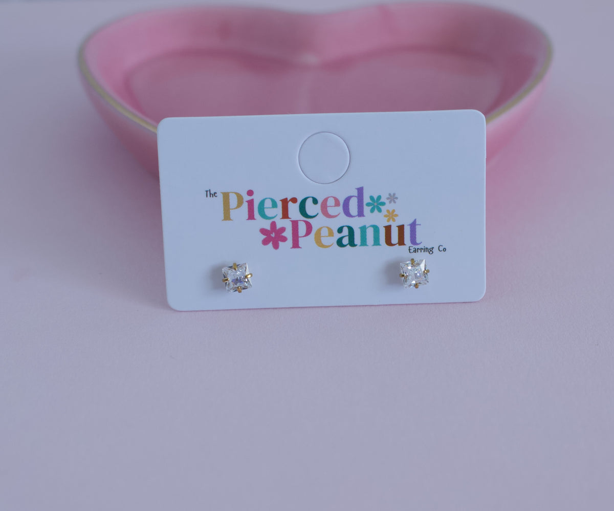 A pair of small silver-toned stud earrings with clear gemstones, displayed on a white branded card that reads 'The Pierced Peanut Earring Co.' against a soft pink background.