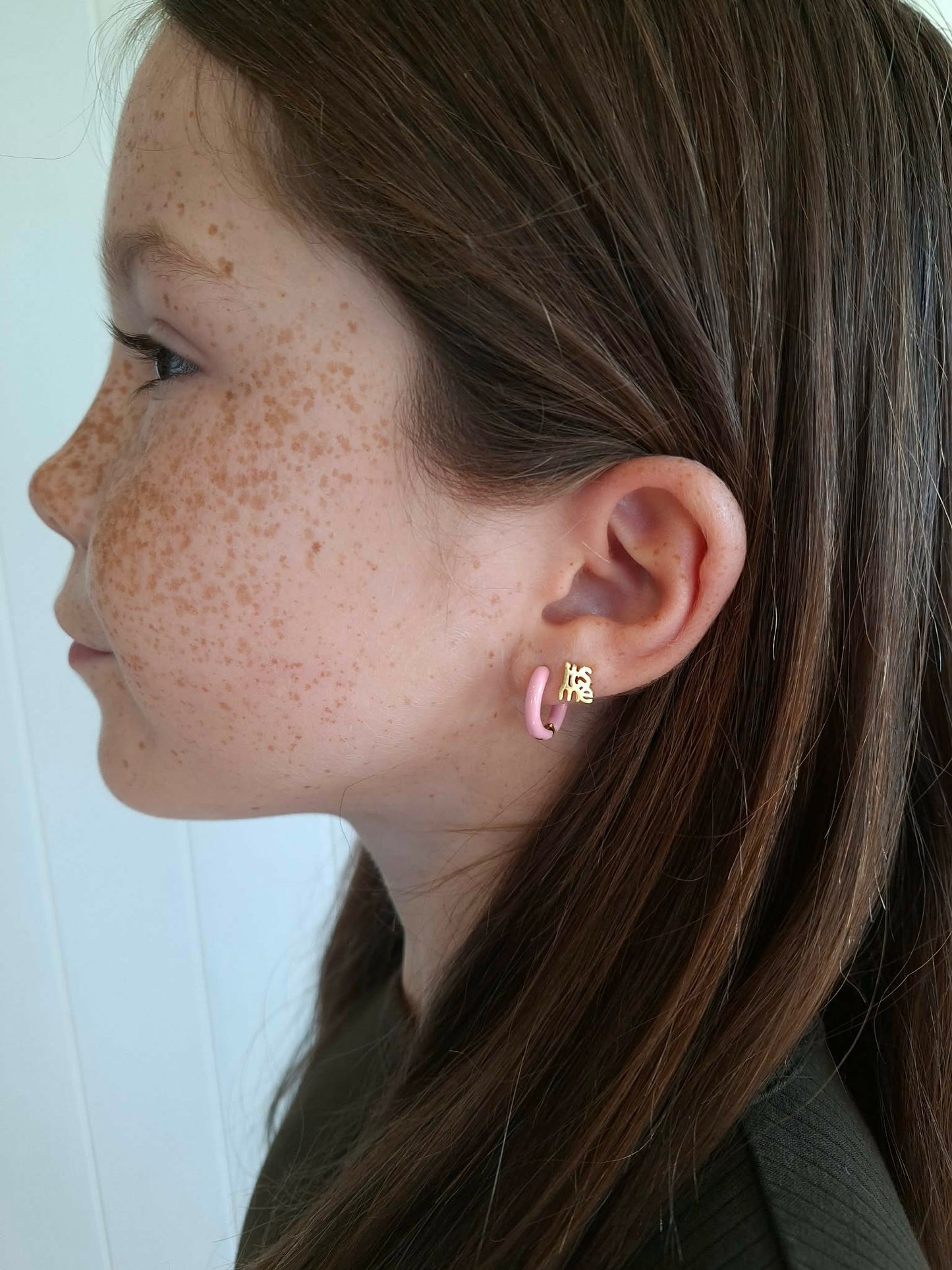 Side view of a girl wearing pastel pink hypoallergenic huggie hoop earrings and a gold screwback stud earring saying "its me" from the Pierced Peanut.