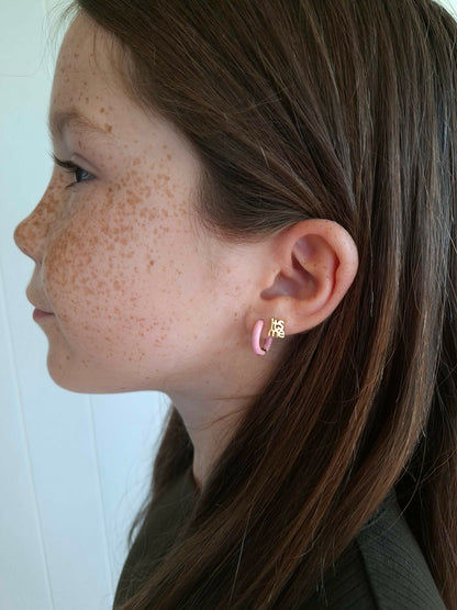 Side view of a girl wearing pastel pink hypoallergenic huggie hoop earrings and a gold screwback stud earring saying "its me" from the Pierced Peanut.