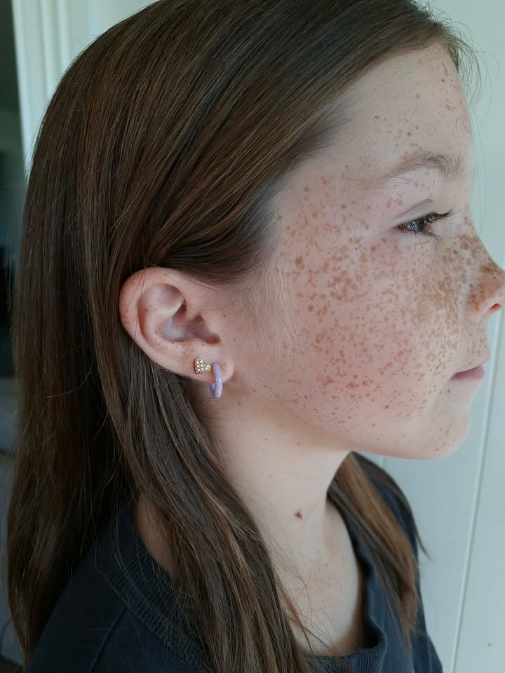 Side view of a child wearing two small hypoallergenic earrings from the pierced peanut. One is a checkered heart screwback stud and the other a pale purple hoop huggie earring.
