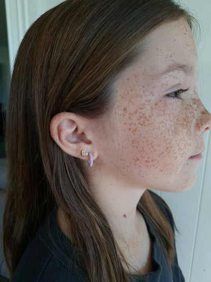 Side view of a child wearing two small hypoallergenic earrings from the pierced peanut. One is a checkered heart screwback stud and the other a pale purple hoop huggie earring.