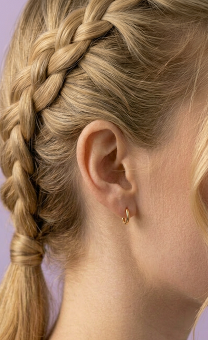 Close-up of a girl with braided hair wearing a gold hypoallergenic huggie earring in one ear from the Pierced Peanut on a light purple background.