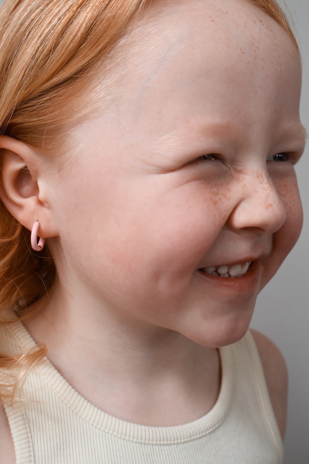 A close-up portrait of a smiling young girl with red hair and freckles, wearing a small pink hoop earring, against a plain light gray background. Huggie earrings are from The Pierced Peanut in NZ.