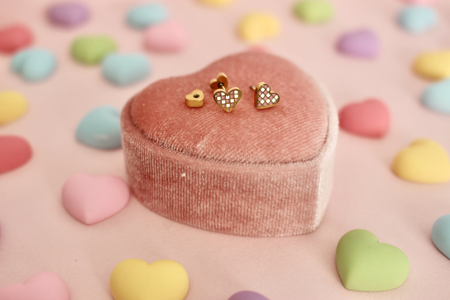 A pair of kids small hypoallergenic gold heart, screwback- stud earrings with white and pink enamel checks, displayed on a pink velvet heart-shaped jewellery box against a soft pink background. The brand of the Earrings in the Pierced Peanut Earring Co in New Zealand.