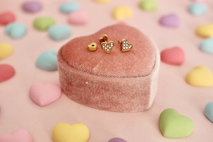 A pair of kids small hypoallergenic gold heart, screwback- stud earrings with white and pink enamel checks, displayed on a pink velvet heart-shaped jewellery box against a soft pink background. The brand of the Earrings in the Pierced Peanut Earring Co in New Zealand.