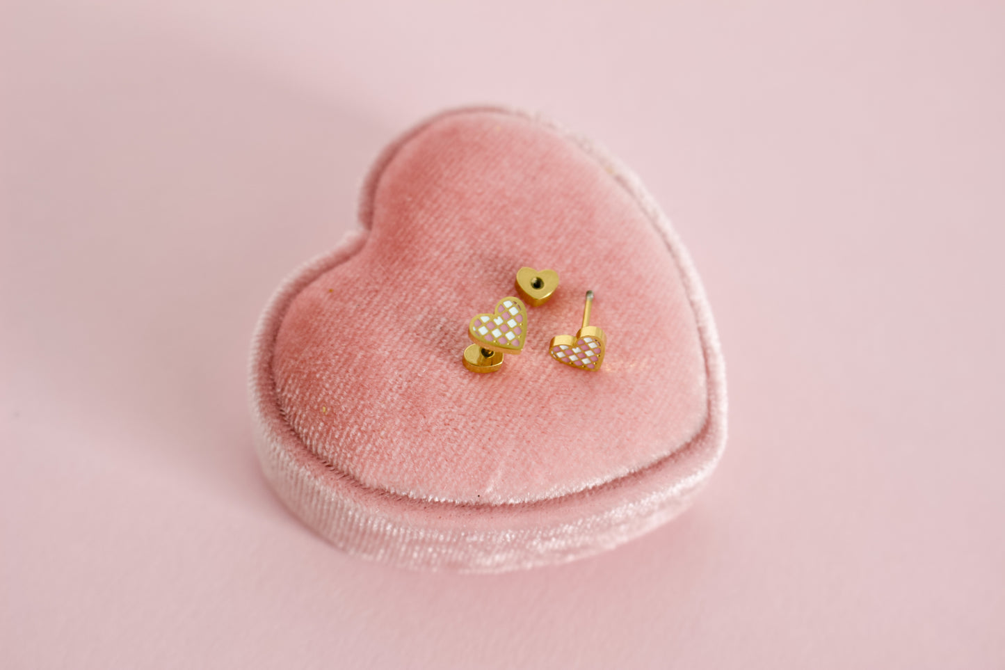 A pair of kids small hypoallergenic gold heart, screwback- stud earrings with white and pink enamel checks, displayed on a pink velvet heart-shaped jewellery box against a soft pink background. The brand of the Earrings in the Pierced Peanut Earring Co in New Zealand.