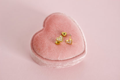 A pair of kids small hypoallergenic gold heart, screwback- stud earrings with white and pink enamel checks, displayed on a pink velvet heart-shaped jewellery box against a soft pink background. The brand of the Earrings in the Pierced Peanut Earring Co in New Zealand.