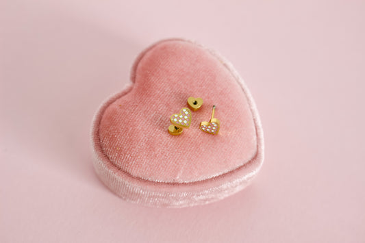 A pair of kids small hypoallergenic gold heart, screwback- stud earrings with white and pink enamel checks, displayed on a pink velvet heart-shaped jewellery box against a soft pink background. The brand of the Earrings in the Pierced Peanut Earring Co in New Zealand.