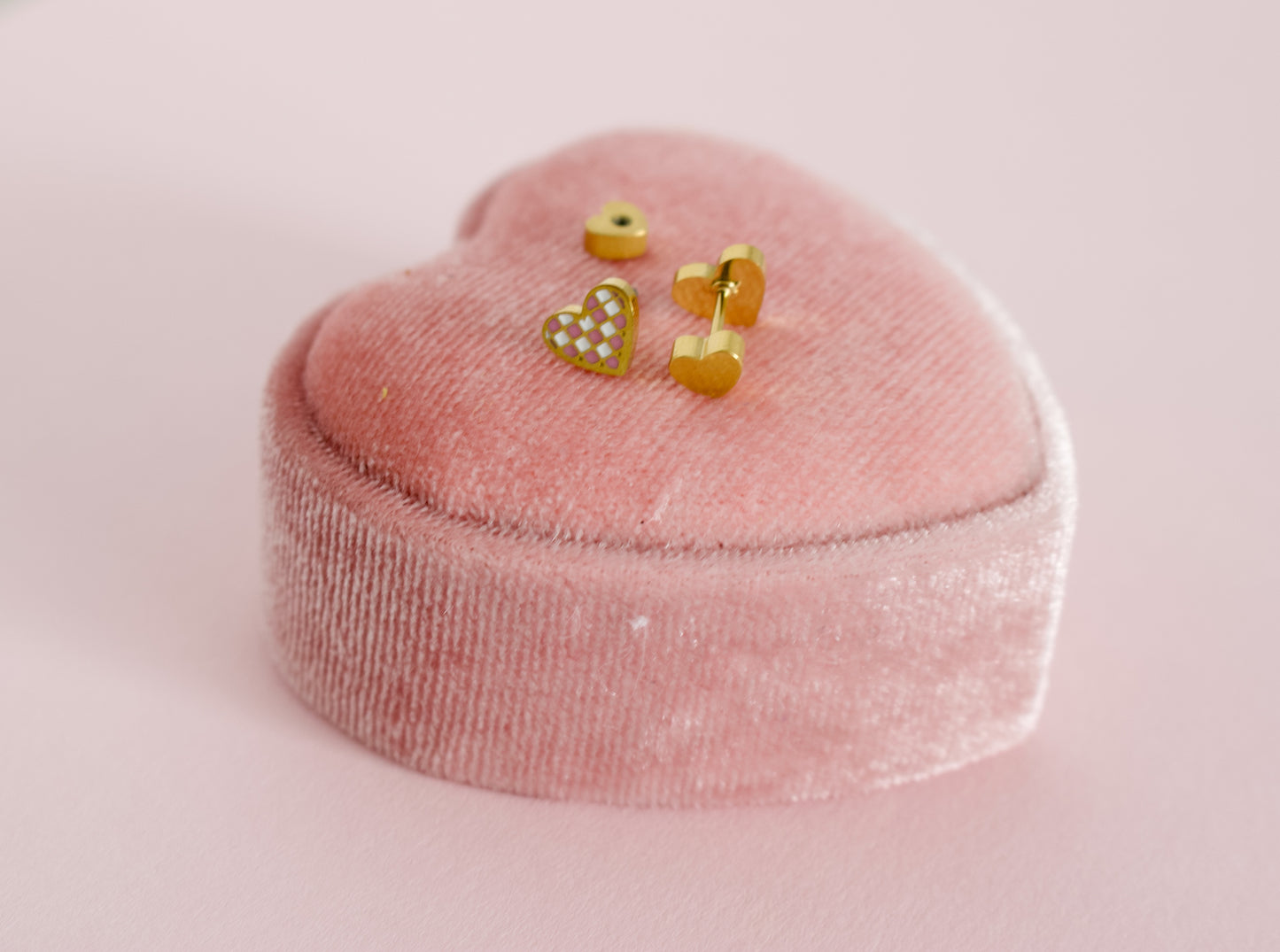 A pair of kids small hypoallergenic gold heart, screwback- stud earrings with white and pink enamel checks, displayed on a pink velvet heart-shaped jewellery box against a soft pink background. The brand of the Earrings in the Pierced Peanut Earring Co in New Zealand.