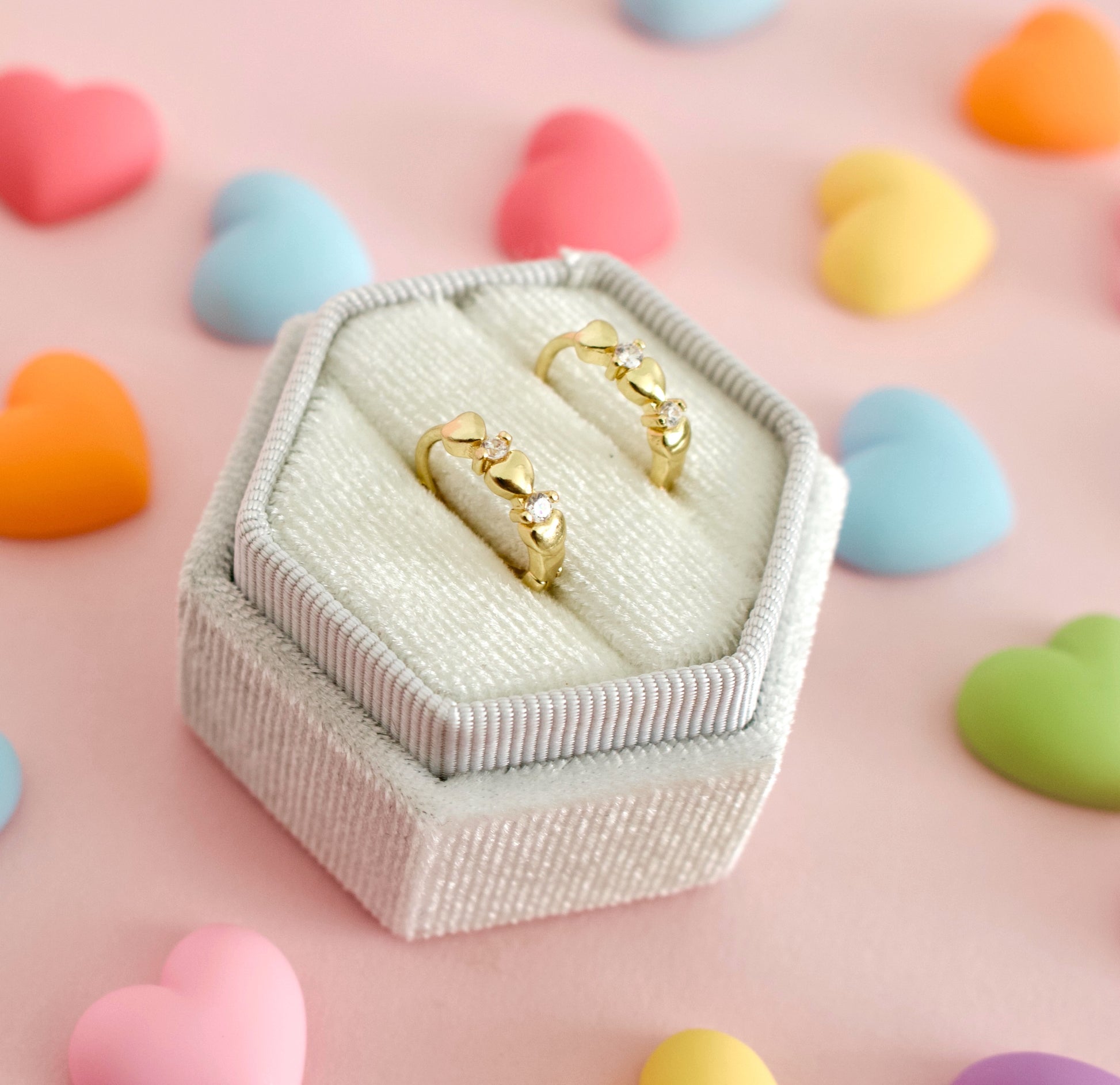 Two gold hypoallergenic huggie hoop earrings with small heart charms displayed in a small white textured ring box, set against a pink background with scattered pastel heart decorations.