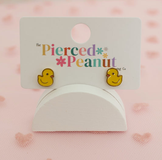 A pair of yellow rubber duck stud earrings displayed on a white card from 'The Pierced Peanut Co', resting on a white stand against a pink background with subtle heart patterns. Hypoallergenic kids screwback earrings
