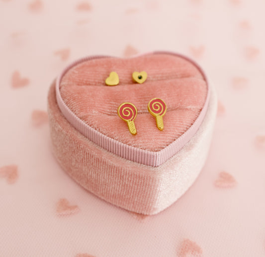 Gold lollipop hypoallergenic stud screwback earrings from The Pierced Peanut displayed on a pink heart-shaped velvet jewellery box against a pink background with heart patterns.
