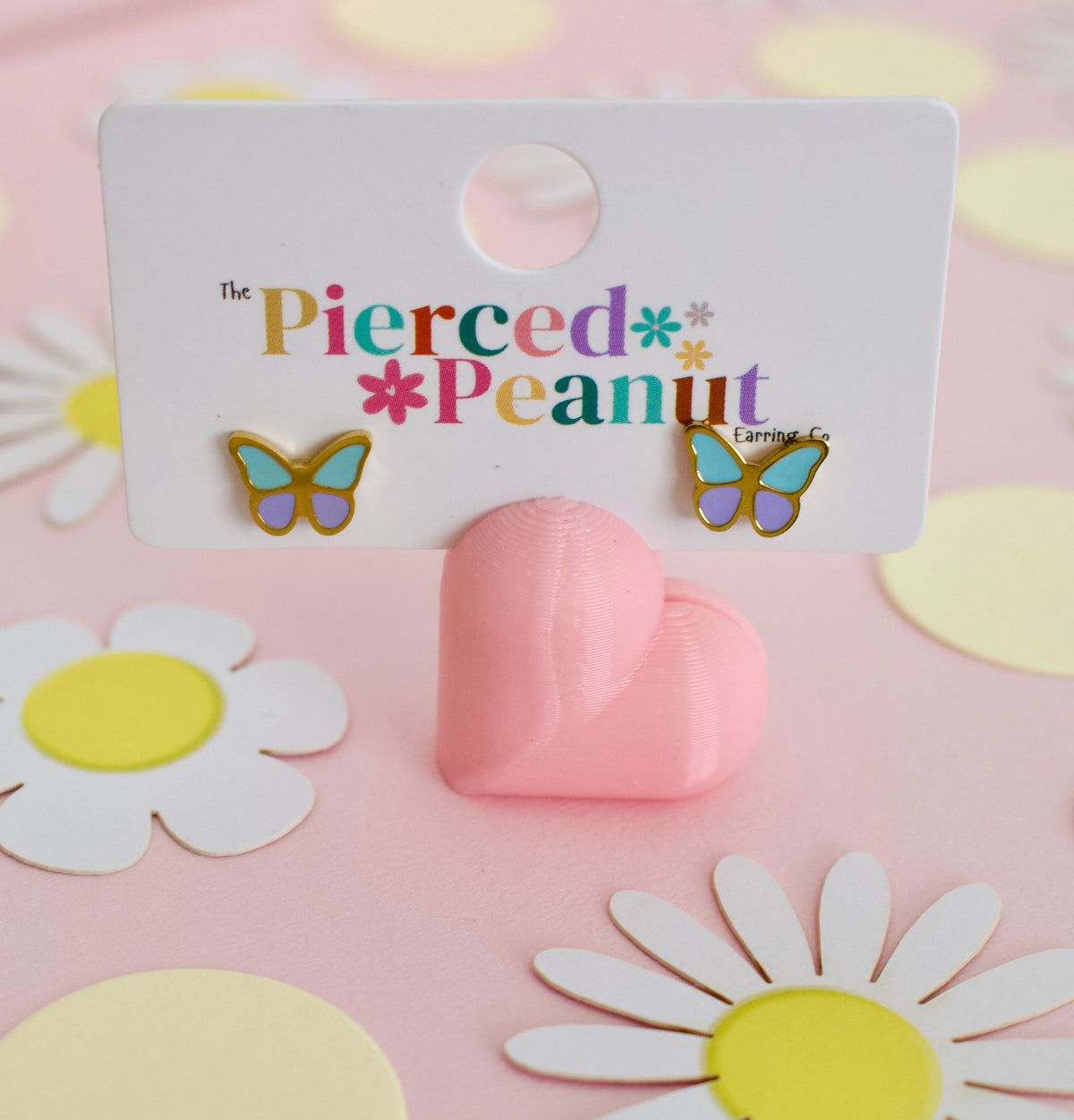 A pair of hypoallergenic butterfly stud screwback earrings from the Pierced Peanut with gold-tone edges, light blue wings, and purple centers, displayed on a white branded backing card that reads 'The Pierced Peanut.' The card is propped by a pink heart-shaped stand on a pink surface with daisy cutouts in the background.