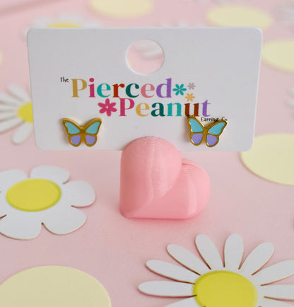 A pair of hypoallergenic butterfly stud screwback earrings from the Pierced Peanut with gold-tone edges, light blue wings, and purple centers, displayed on a white branded backing card that reads 'The Pierced Peanut.' The card is propped by a pink heart-shaped stand on a pink surface with daisy cutouts in the background.