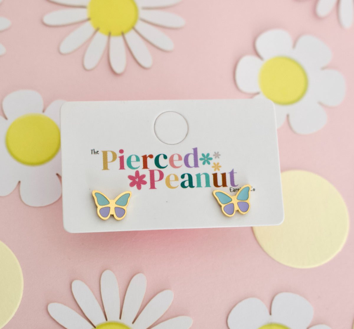 A pair of hypoallergenic butterfly screwback stud earrings with gold-tone edges, pastel purple centers, and mint green wings, displayed on a white branded card that reads 'The Pierced Peanut.' The card is placed on a pink background decorated with white daisy cutouts and yellow circles.