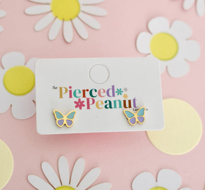 A pair of hypoallergenic butterfly screwback stud earrings with gold-tone edges, pastel purple centers, and mint green wings, displayed on a white branded card that reads 'The Pierced Peanut.' The card is placed on a pink background decorated with white daisy cutouts and yellow circles.