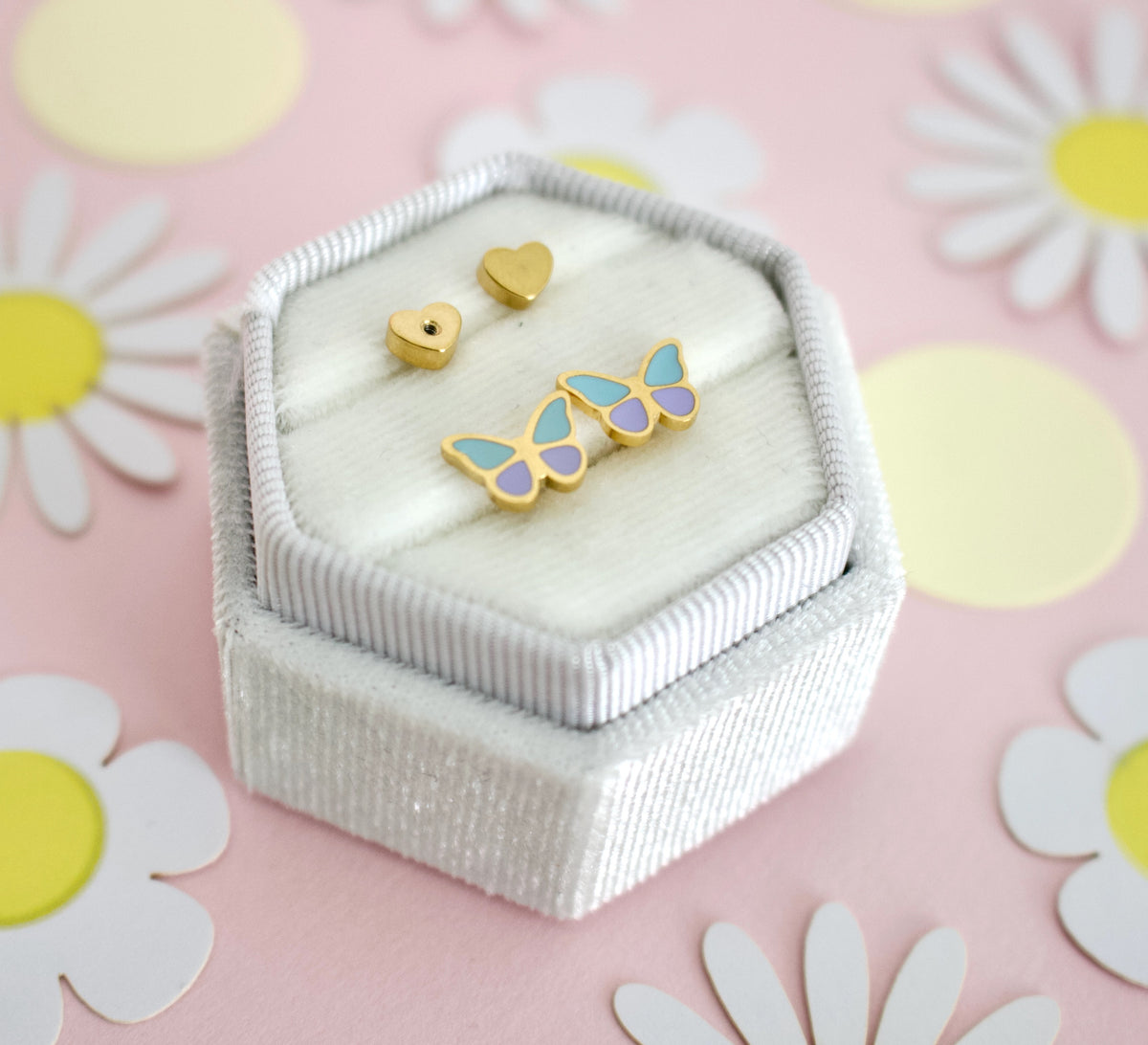A pair of hypoallergenic gold butterfly screwback stud earrings from the Pierced Peanut with light blue and lavender enamel wings, displayed on a white ribbed jewelry box next to two small gold heart studs, set against a pink background with white and yellow daisy decorations.