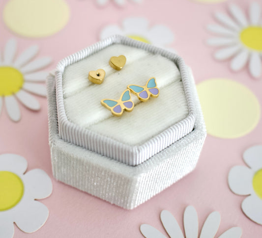 A pair of hypoallergenic gold butterfly screwback stud earrings from the Pierced Peanut with light blue and lavender enamel wings, displayed on a white ribbed jewelry box next to two small gold heart studs, set against a pink background with white and yellow daisy decorations.