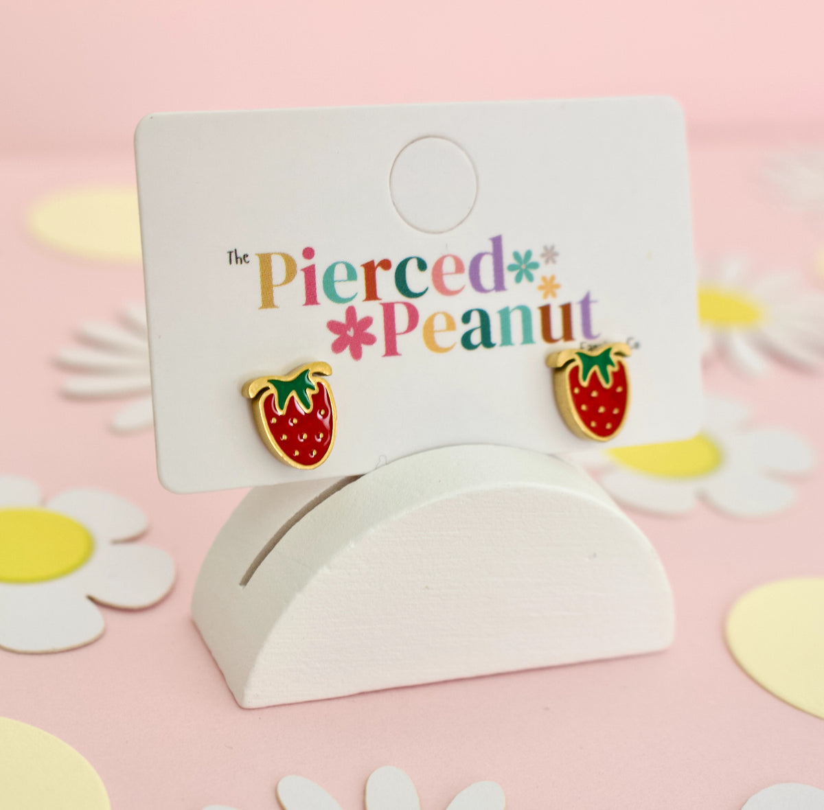 A pair of hypoallergenic strawberry-shaped screwback stud earrings displayed on a white card stand. The earrings are red with green leaf tops and gold-tone edges, mounted on a white card printed with the brand name 'The Pierced Peanut'.