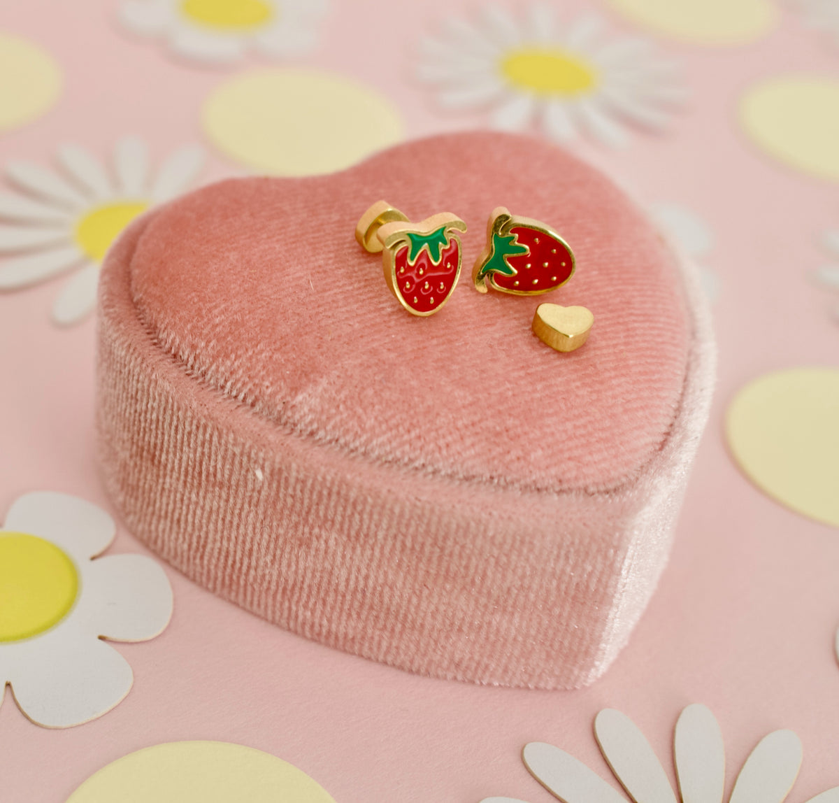 A pair of hypoallergenic strawberry screwback stud earrings from the Pierced PEanut with gold-plated edges, red enamel, and green leaf tops, displayed on a pink heart-shaped velvet jewelry box against a pink background decorated with white daisies and yellow circles.