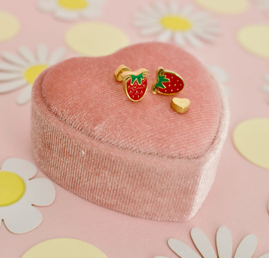A pair of hypoallergenic strawberry screwback stud earrings from the Pierced PEanut with gold-plated edges, red enamel, and green leaf tops, displayed on a pink heart-shaped velvet jewelry box against a pink background decorated with white daisies and yellow circles.