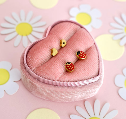 A pair of small hypoallergenic ladybug heart screwback stud earrings from the Pierced Peanut displayed on a pink heart-shaped jewelry box, set against a pink background with white and yellow daisy cutouts.