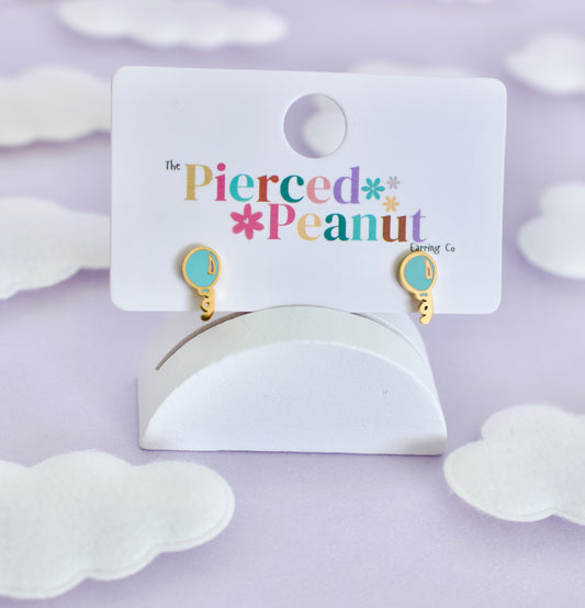 A pair of hypoallergenic gold and light blue balloon screwback stud earrings, displayed on a white card stand that reads 'The Pierced Peanut Earring Co'. The stand is set against a soft lavender background decorated with white cloud cutouts.