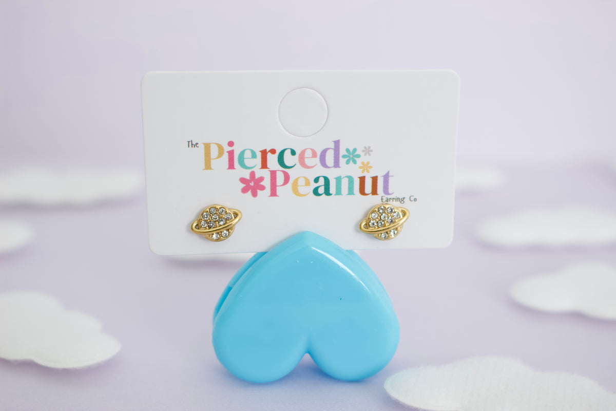 A pair of hypoallergenic gold-toned space-themed screwback stud earrings with clear rhinestones, displayed on a white card labeled 'The Pierced Peanut Living Co'. A light blue heart-shaped prop sits in front of the card against a soft lavender background with white cloud cutouts.
