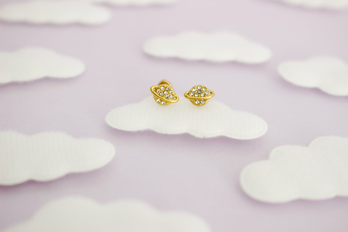 A pair of hypoallergenic small gold space-themed screwback stud earrings with tiny clear gemstones, displayed on a white cloud-shaped prop against a soft lavender background with more cloud cutouts.
