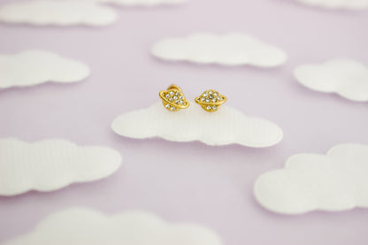 A pair of hypoallergenic small gold space-themed screwback stud earrings with tiny clear gemstones, displayed on a white cloud-shaped prop against a soft lavender background with more cloud cutouts.