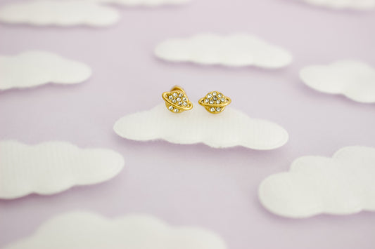 A pair of hypoallergenic small gold space-themed screwback stud earrings with tiny clear gemstones, displayed on a white cloud-shaped prop against a soft lavender background with more cloud cutouts.