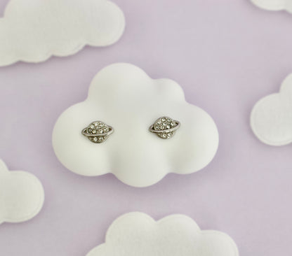 A pair of silver-tone hypoallergenic screwback stud planet shaped earrings from the Pierced Peanut. Displayed on a white cloud-shaped stand against a pale lavender background with additional cloud-shaped props.