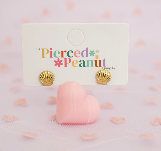 A pair of hypoallergenic gold shell screwback stud earrings displayed on a white card labeled 'The Pierced Peanut Loring Co', with a pink heart-shaped prop in front on a soft pink background.