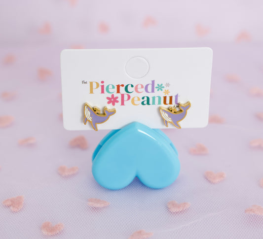 A pair of small hypoallergenic gold-tone and lavender whale screwback stud earrings displayed on a white branded card, with a glossy light blue heart-shaped stand in front. The card reads 'the Pierced * Peanut'.