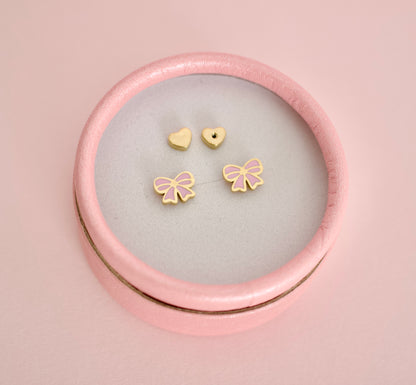 A set of small hypoallergenic gold-tone screwback stud earrings displayed inside a round pink presentation box on a soft pink background. The earrings include two heart-shaped screwbacks and two bow-shaped studs with pink enamel detailing.