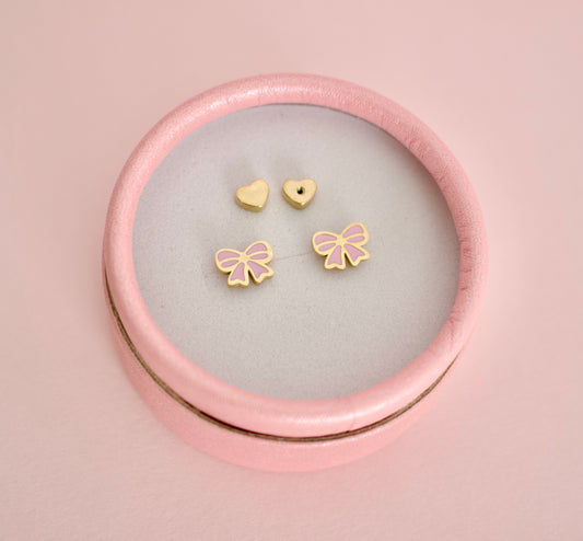 A set of small hypoallergenic gold-tone screwback stud earrings displayed inside a round pink presentation box on a soft pink background. The earrings include two heart-shaped screwbacks and two bow-shaped studs with pink enamel detailing.