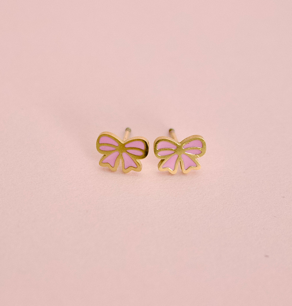 A pair of small bow-shaped stud earrings with gold-tone outlines and light pink enamel fillings, displayed on a soft pink background.