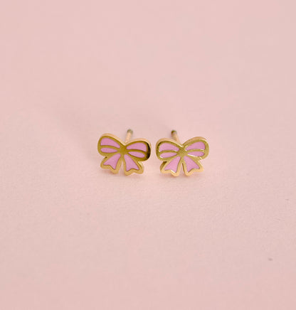 A pair of small bow-shaped stud earrings with gold-tone outlines and light pink enamel fillings, displayed on a soft pink background.