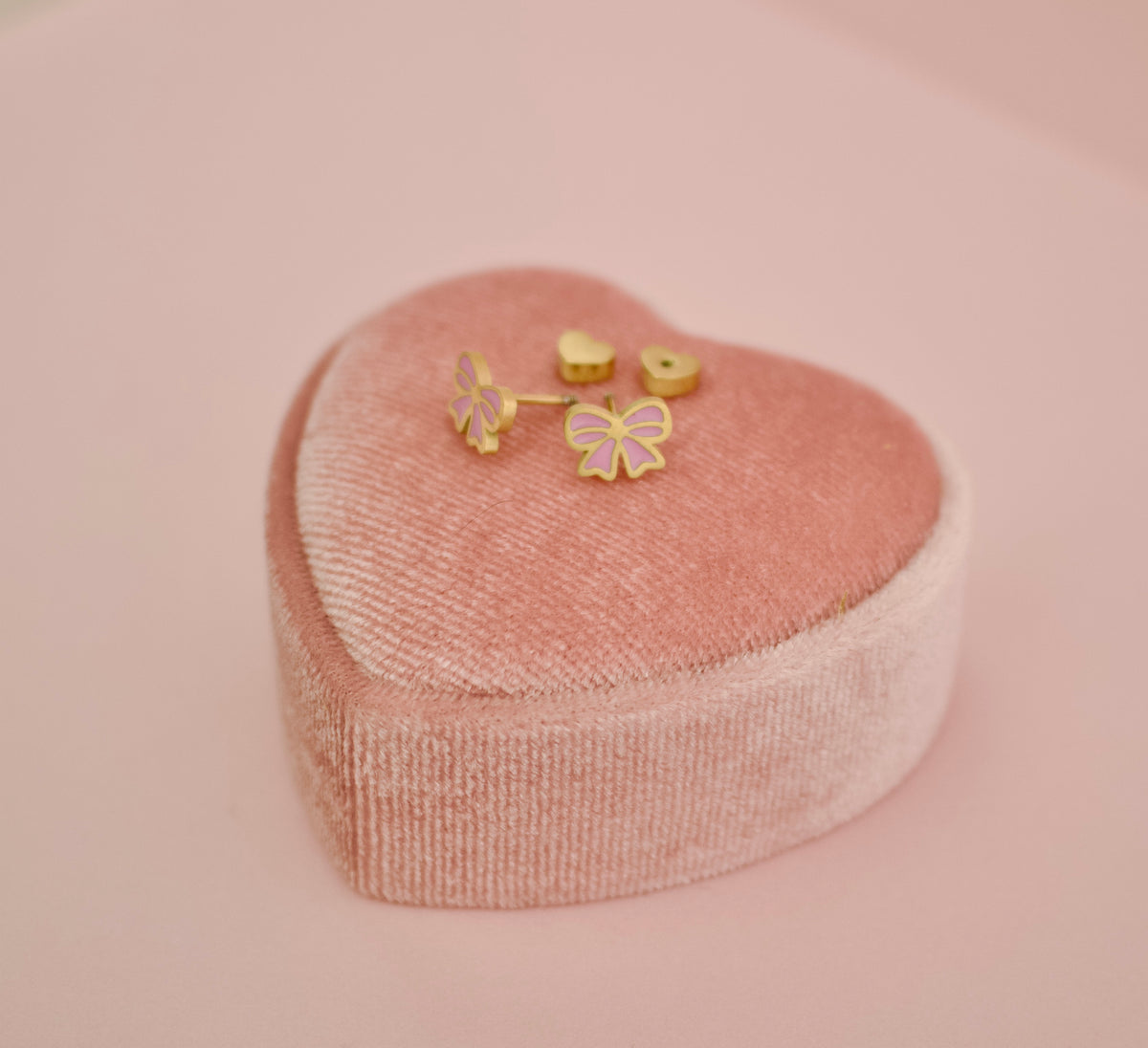 A set of small hypoallergenic gold-tone screwback stud earrings displayed inside a heart shaped pink presentation box on a soft pink background. The earrings include two heart-shaped screwbacks and two bow-shaped studs with pink enamel detailing.