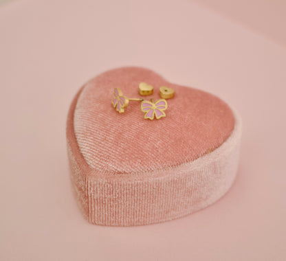 A set of small hypoallergenic gold-tone screwback stud earrings displayed inside a heart shaped pink presentation box on a soft pink background. The earrings include two heart-shaped screwbacks and two bow-shaped studs with pink enamel detailing.