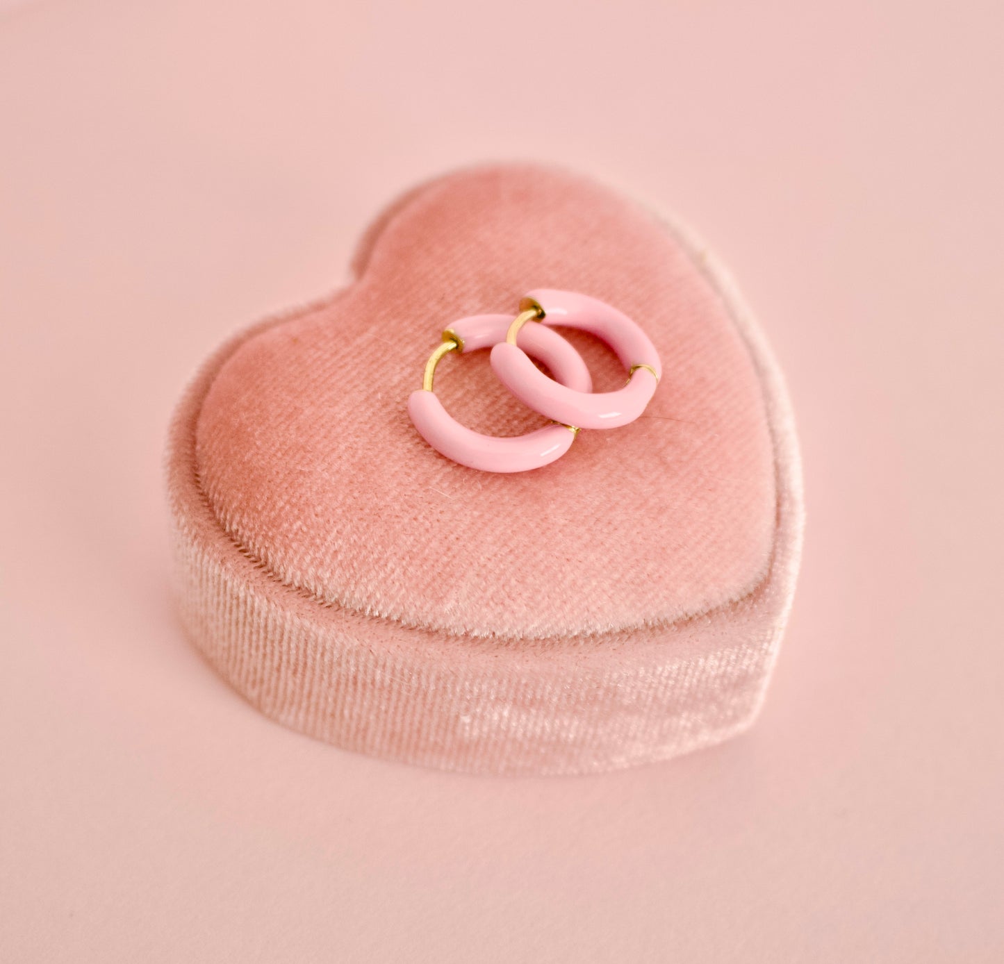 A pair of hypoallergenic pink enamel hoop earrings with gold-tone metal wire loops, displayed on a pink velvet heart-shaped ring box against a solid pink background from the Pierced Peanut earring co.