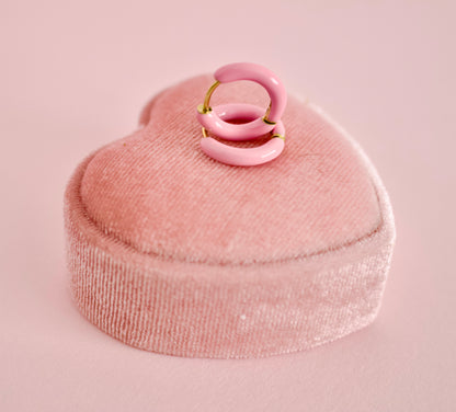 A pair of hypoallergenic pink enamel hoop earrings with gold-tone metal wire loops, displayed on a pink velvet heart-shaped ring box against a solid pink background from the Pierced Peanut earring co.