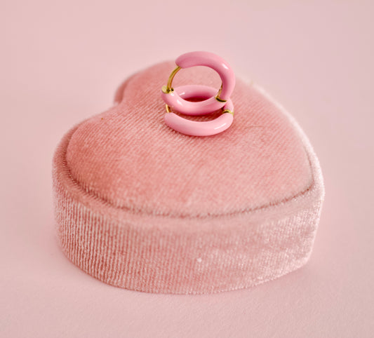 A pair of hypoallergenic pink enamel hoop earrings with gold-tone metal wire loops, displayed on a pink velvet heart-shaped ring box against a solid pink background from the Pierced Peanut earring co.