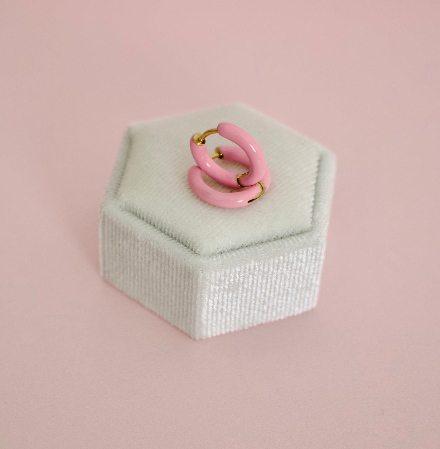 A pair of hypoallergenic pink enamel hoop earrings with gold-tone metal wire loops, displayed on a white velvet ring box against a solid pink background from the Pierced Peanut earring co.